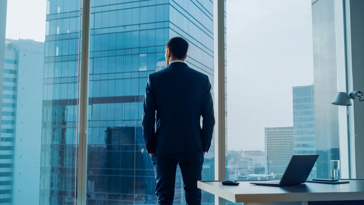 Back View of the Thoughtful Businessman wearing a Suit Standing in His Office, Hands in Pockets and Contemplating Next Big Business Deal, Looking out of the Window. Big City Business District Panoramic Window View.